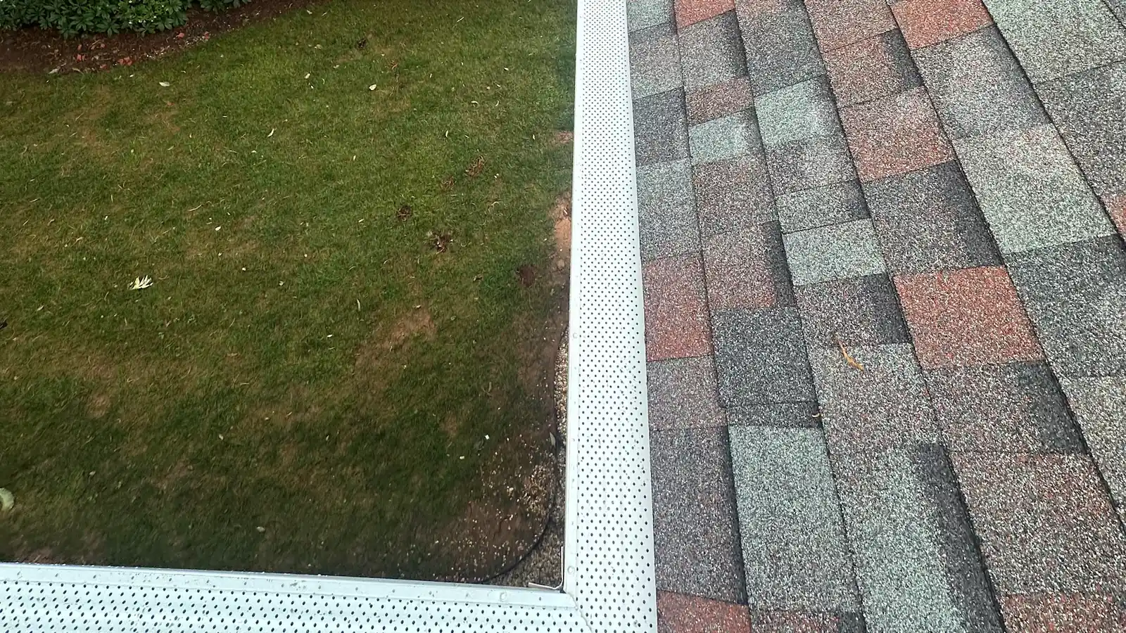 A white perforated gutter guard running along a shingled roof edge above a lawn