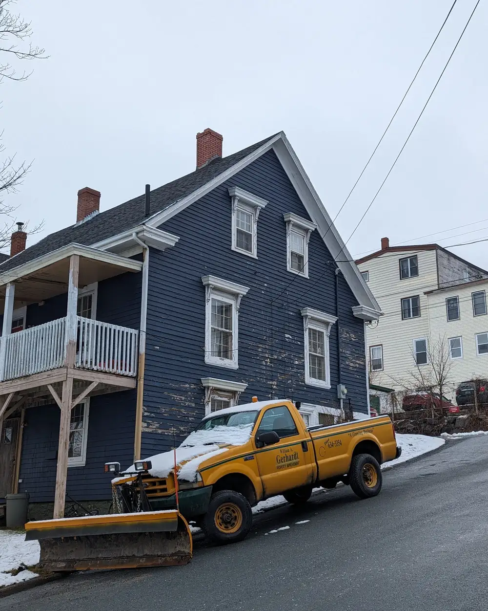 A pickup with a plow blade parked in front of a clapboard house after snow